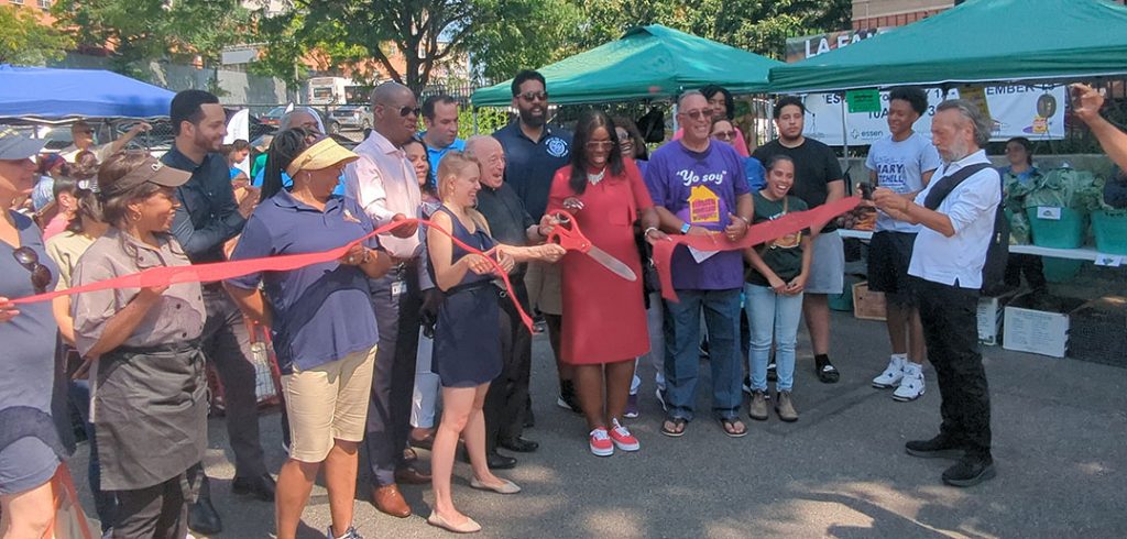 Nine people stand in front of a red ribbon while holding an over-sized pair of scissors