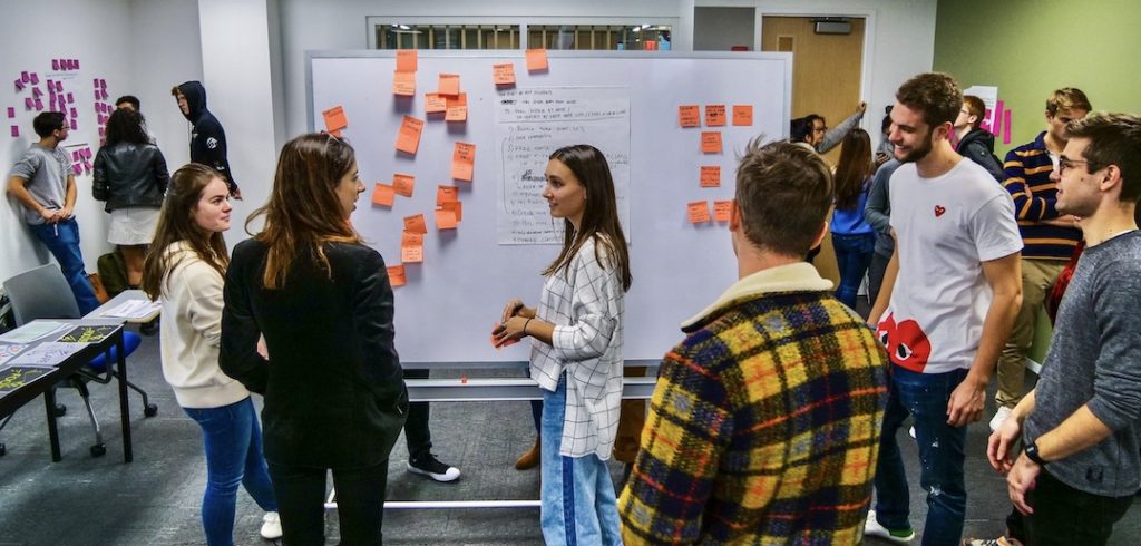 Students stand and speak next to a whiteboard covered with post-its.