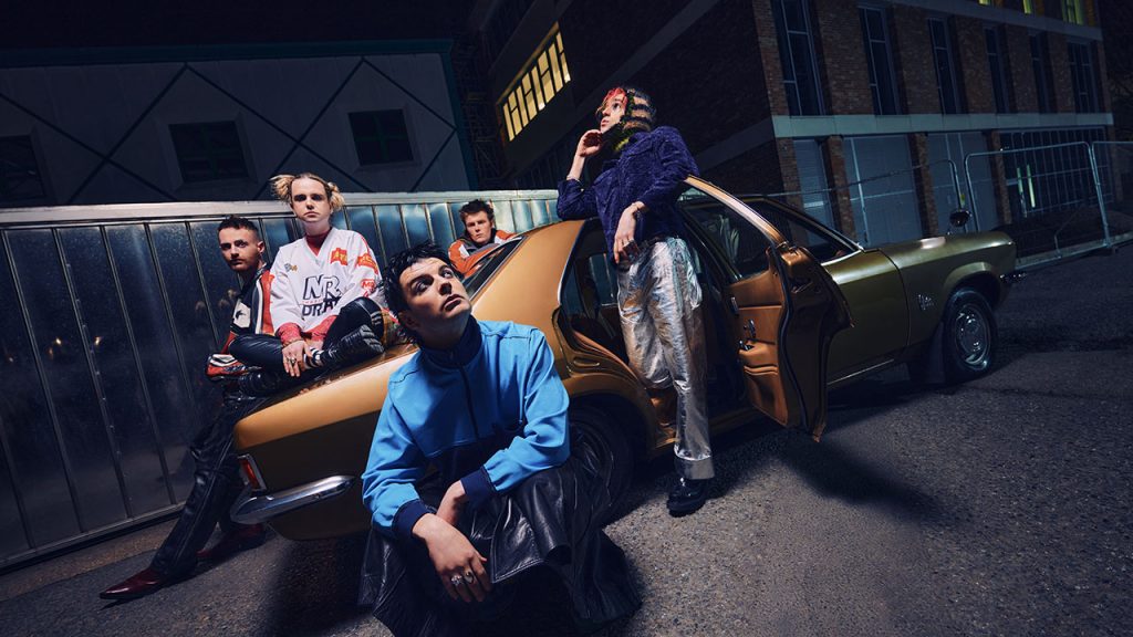 Four people strike poses on a city street, on and around a tan car with the back door open