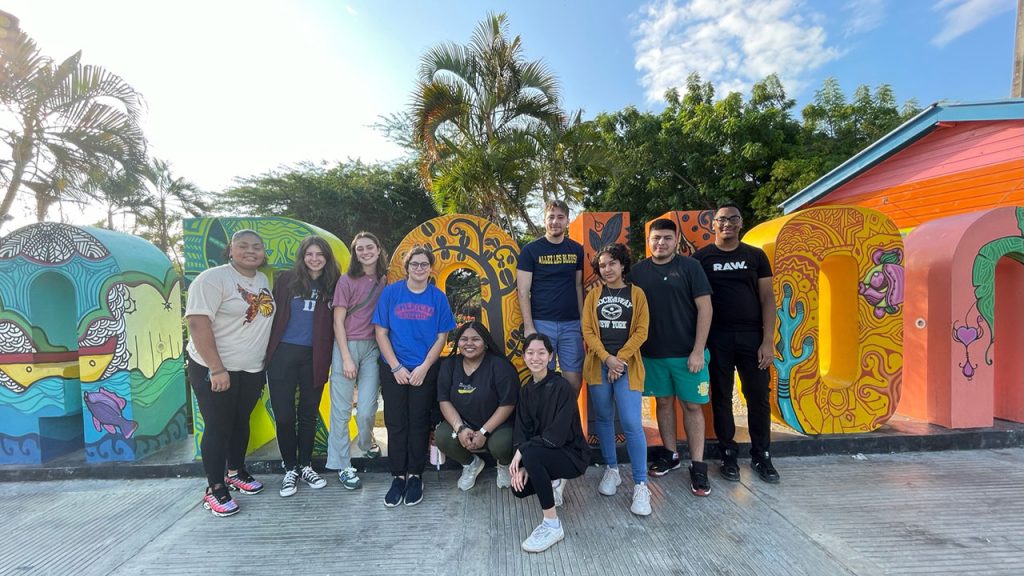 Group photo of female and male presenting individuals in Dominican Republic. They are posing in front of bright colorful life- sized letters, "A" on the left and "O" on the right.