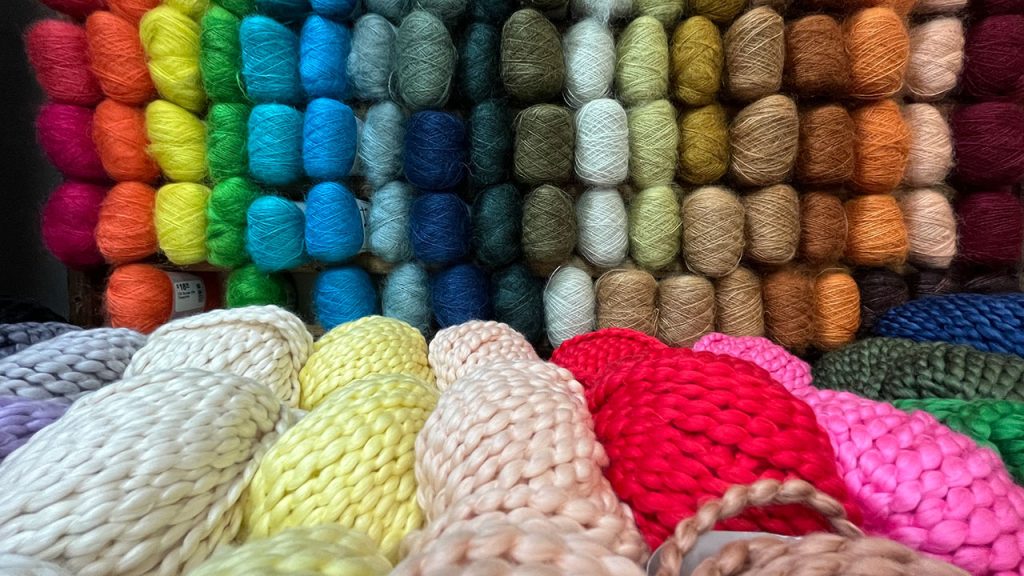 Wall full of multiple colors of yarn, yarn also in the forefront sitting on a table. 
