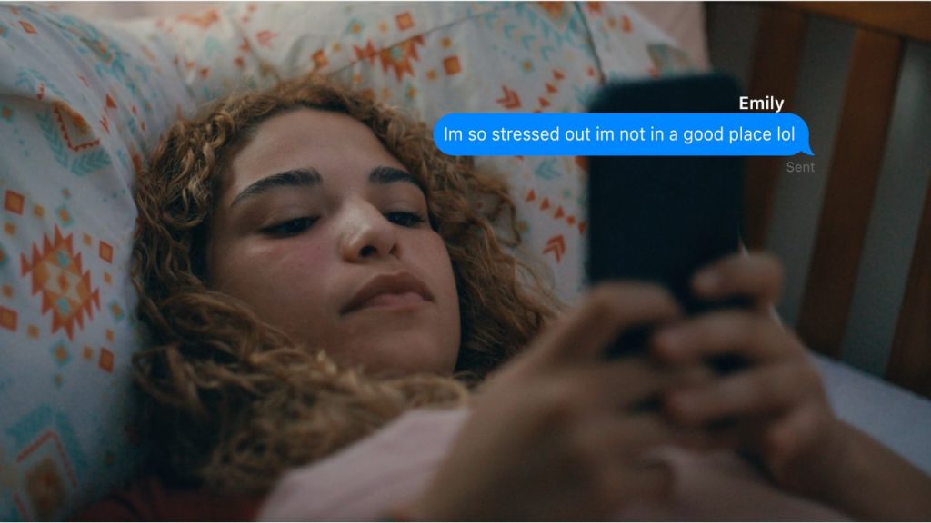 A young girl lies in bed texting on her cellphone with a text bubble reading "I'm so stressed out I'm not in a good place lol," a still shot from the short film "Nuestro Apoyo" by Fordham Faculty Jenn Lilly