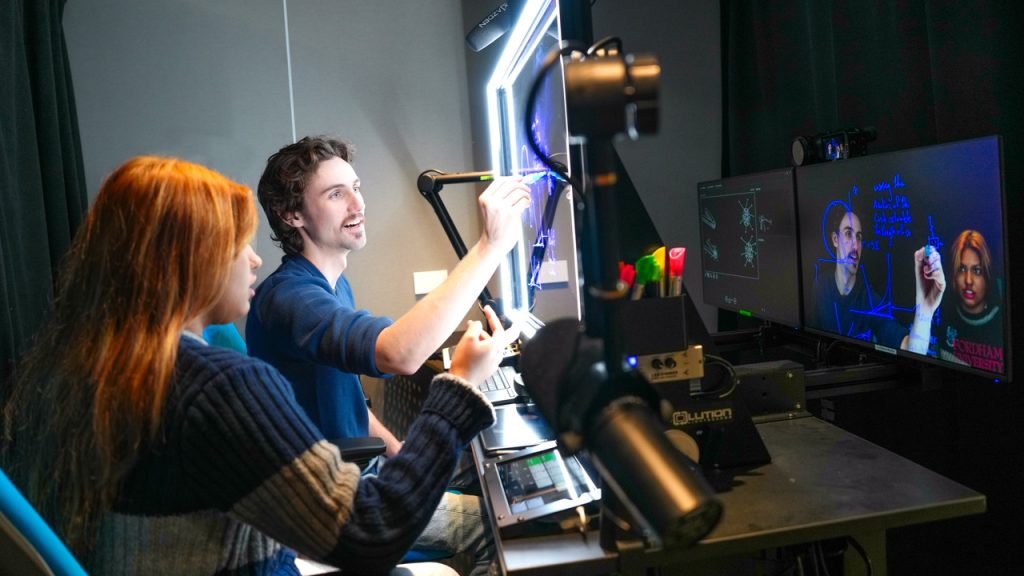 Two Fordham students practice writing on a glass screen in a learning commons "one-button" studio at Fordham.