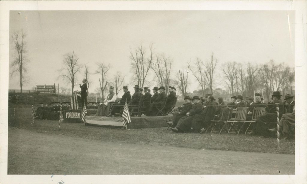 A few dozen priests and dignitaries sit on chairs and a dais set in an open field behind a Fordham banner and two U.S. flags