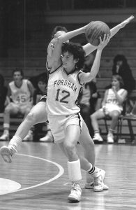 Jeanine “J.J.” Radice, wearing No. 12 for the Fordham women's basketball team, holds the ball above her head and moves around an opponent.