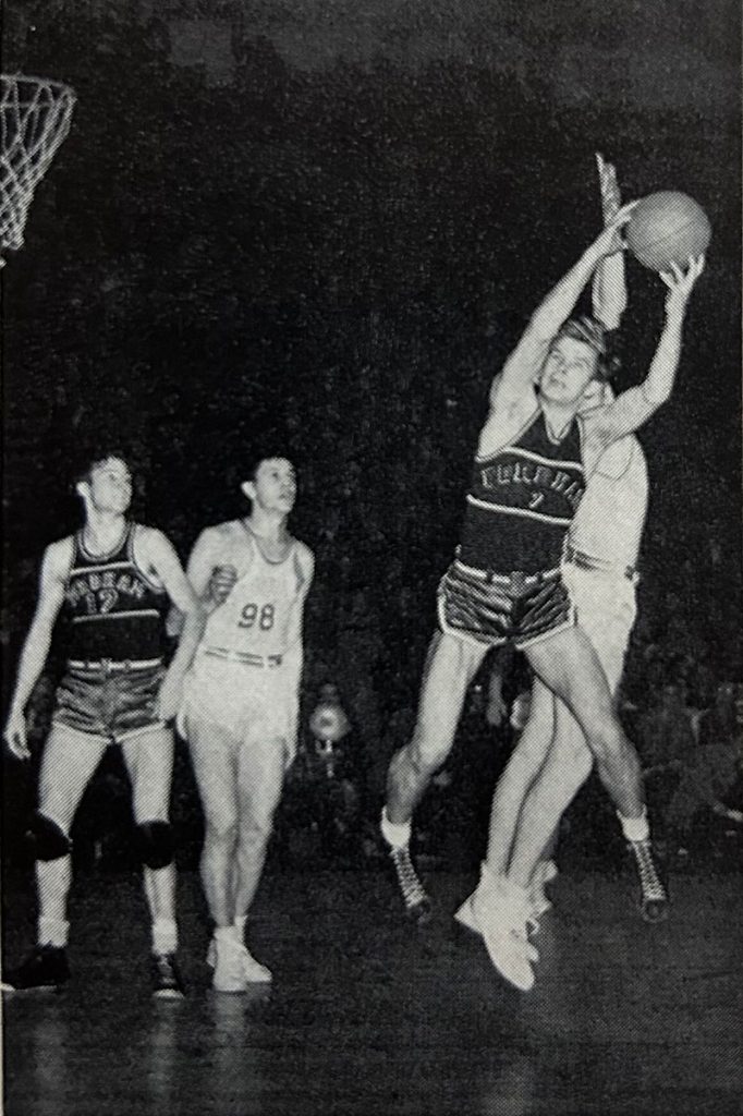 Fordham basketball player Bob Mullens leaps and holds the ball above his head, away from an opponent