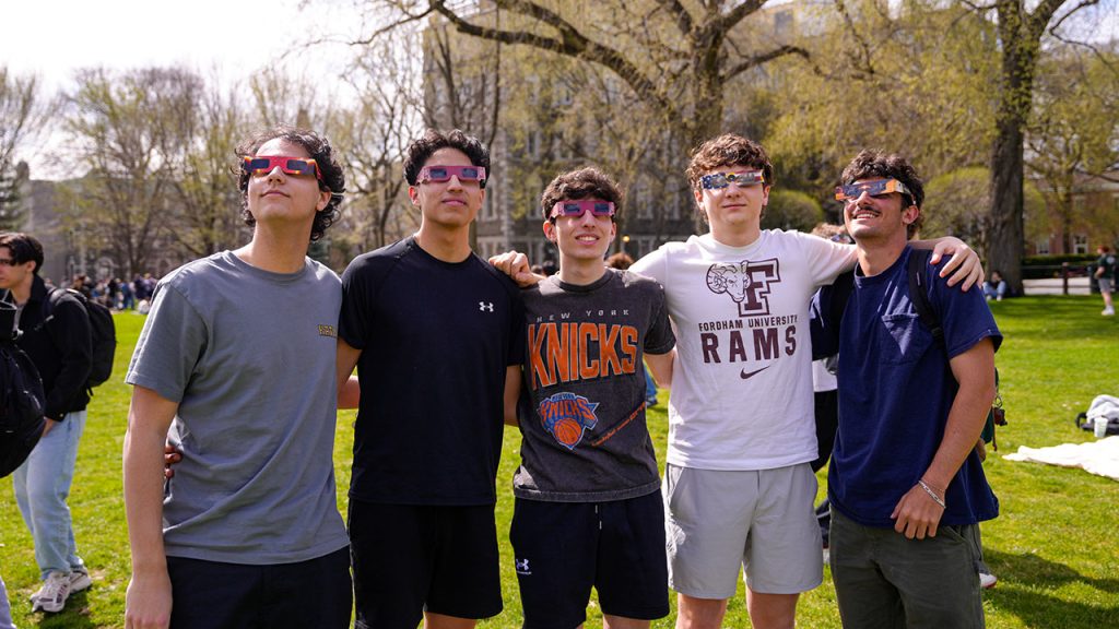 5 male presenting students with arms around each other looking at eclipse, with eclipse glasses on. 