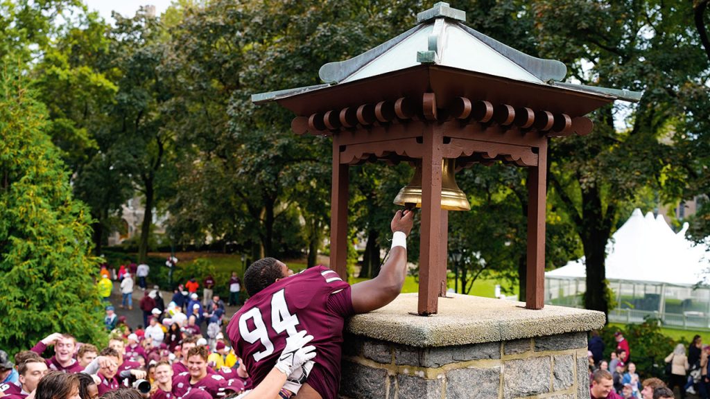 A Fordham football player, boosted by his teammates, rings the Victory Bell amid a crowd of fans and players in front of Fordham's Rose Hill Gym