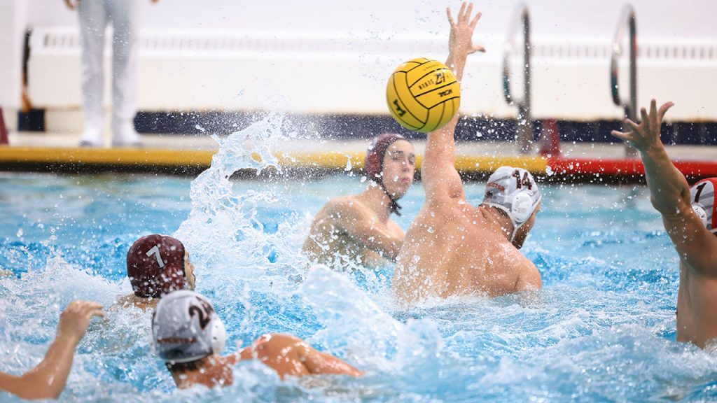 Water polo players in action during a match—one player has just released a shot that evades the outstretched hands of opposing players