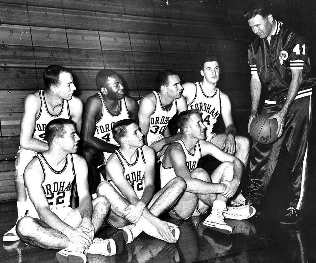 Fordham men's basketball coach Johnny Bach holds a basketball and has the attention of all seven Fordham players crouching and looking up at him in the Rose Hill Gym