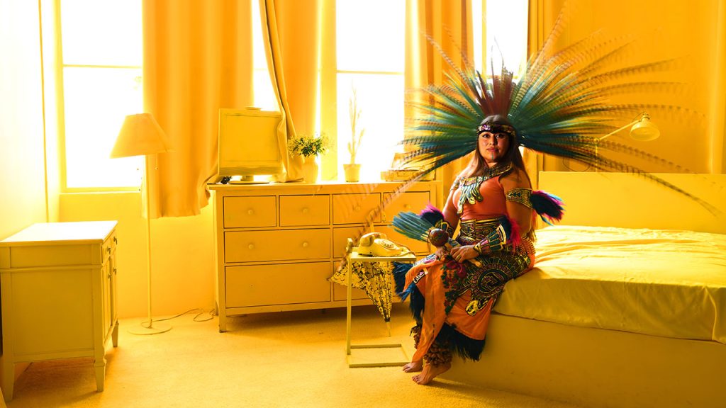 Los Angeles-based folkloric dancer Citlali Arvizu, wearing Aztec headdress and clothing, sits on a yellow bed in a room with yellow curtains and carpeting